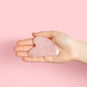 Woman with rose quartz gua sha tool on pink background, closeup